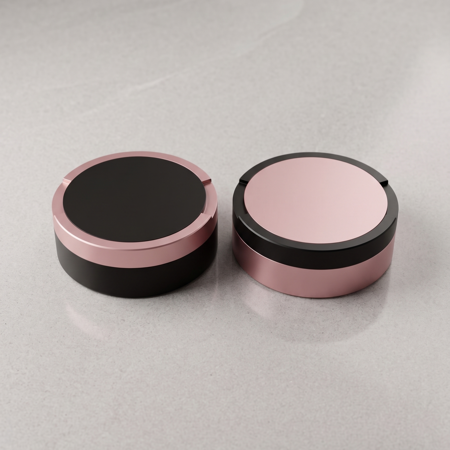 En parbild med två runda metalldosor i storlek large, sida vid sida på en ljusgrå yta. Ena dosa är svart med rosa ring, den andra är rosa med en svart ring. 