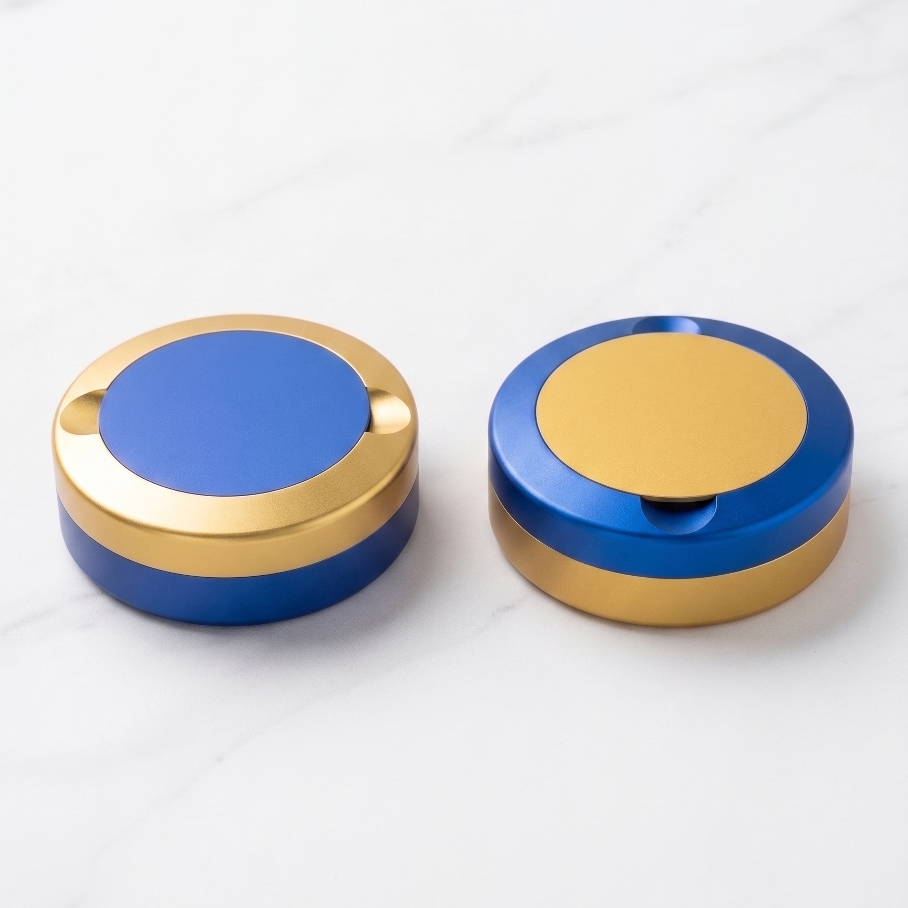 En parbild med två runda metalldosor i storlek slim, sida vid sida på en ljusgrå yta. Ena dosan är blå med guldig ring, den andra är guldig med blå ring. 
