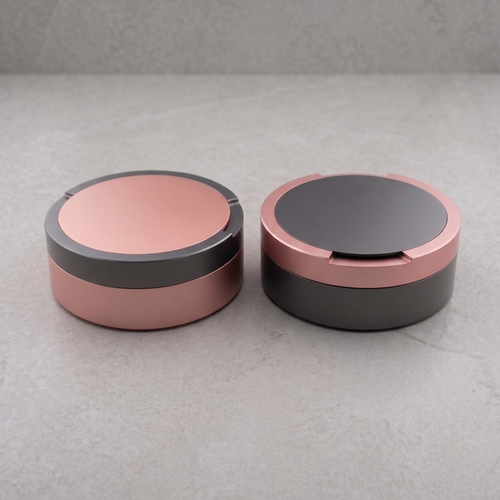 En parbild med två runda metalldosor i storlek large, sida vid sida på en ljusgrå yta. En dosa är grå med rosa ring, den andra är rosa med grå ring. 