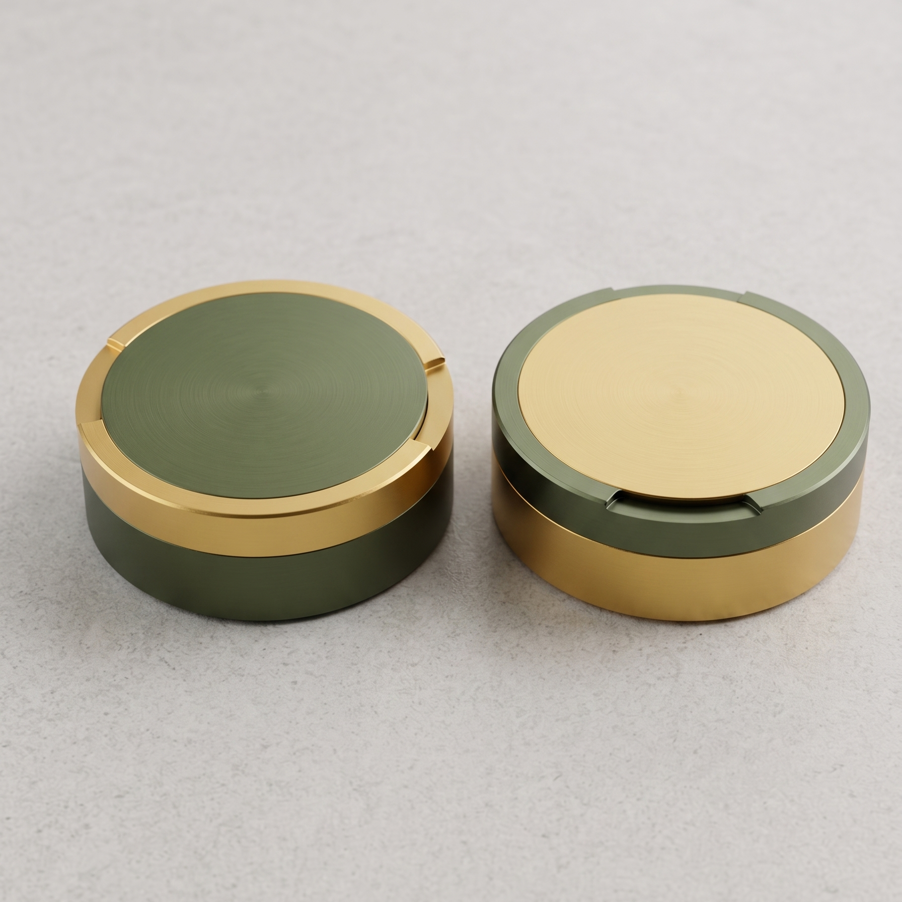 En parbild med två runda metalldosor i storlek large, sida vid sida på en ljusgrå yta. En dosa är olivgrön med guldringar, den andra är guld med en olivgrön ring. 